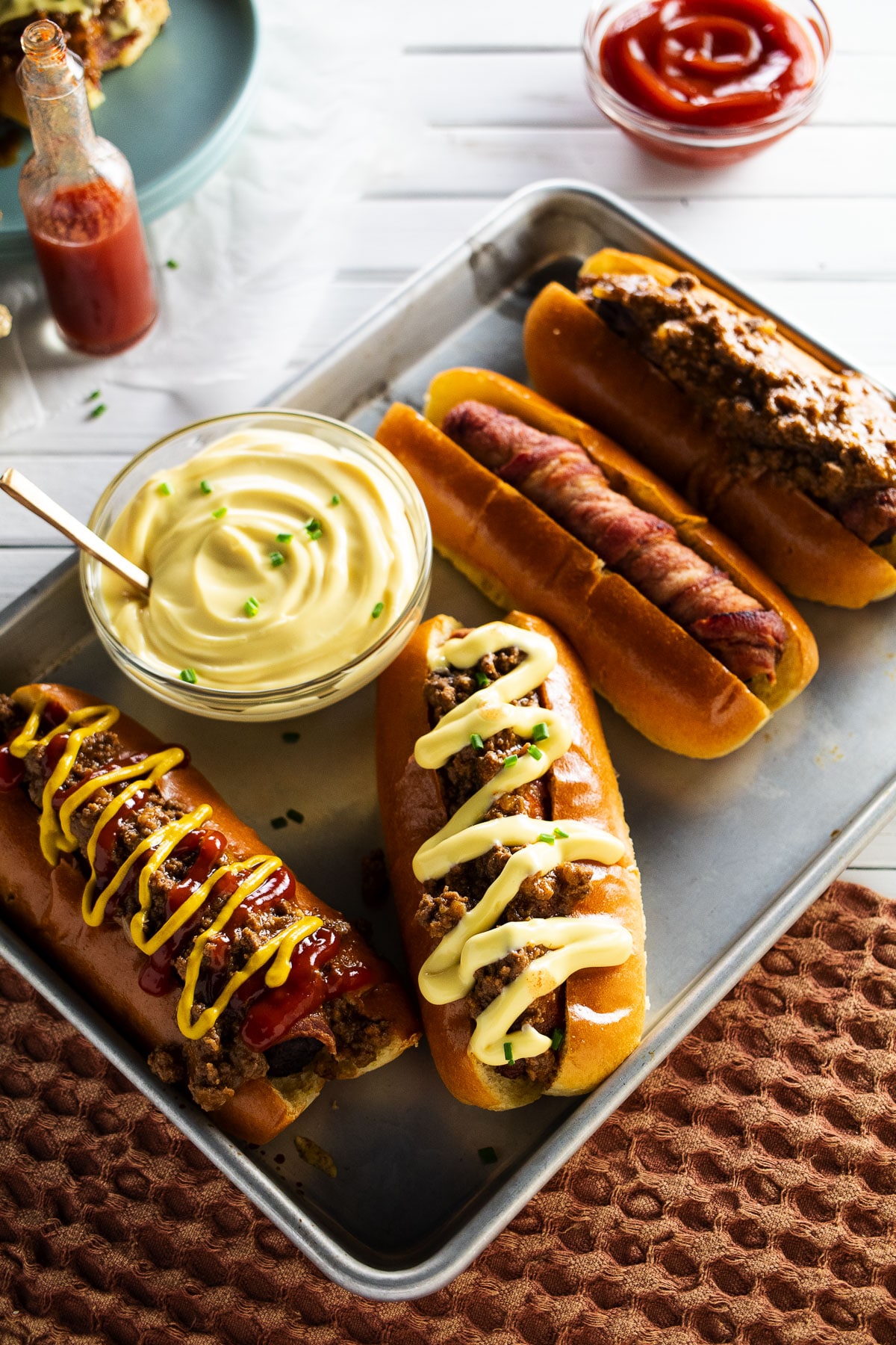 Bacon wrapped chili dogs on pan with nacho cheese, ketchup, and hot suace in the background.