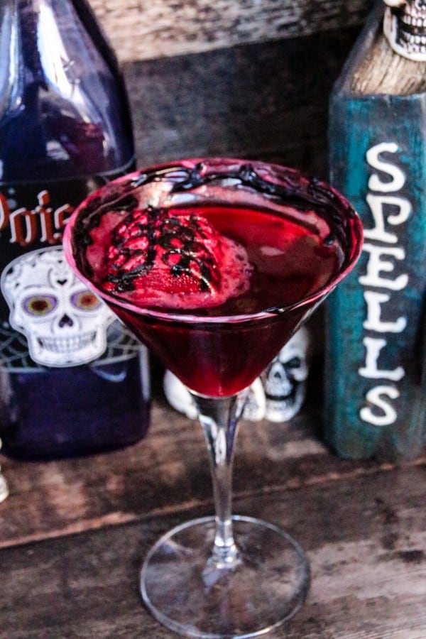 Red mixed drink cocktail in a glass with Halloween decorations in the background.