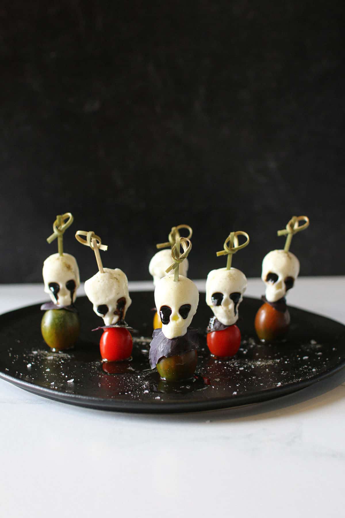 Halloween Caprese skewers with mozzarella skulls, tomatoes, and purple basil on a black plate with black background.