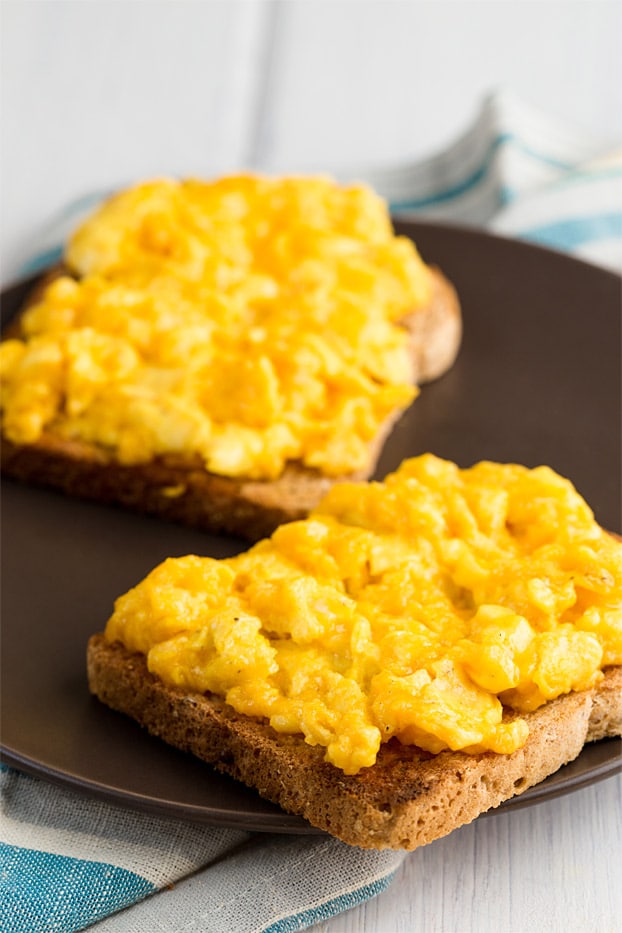 Perfect scrambled eggs with cheese on toast on brown plate and napkin in the background.