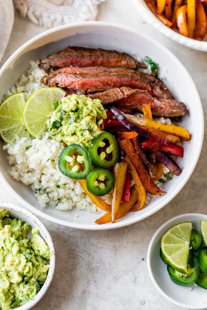 Steak fajita bowl with steak multicolored bell peppers, guacamole, lime wedges in white bowl.