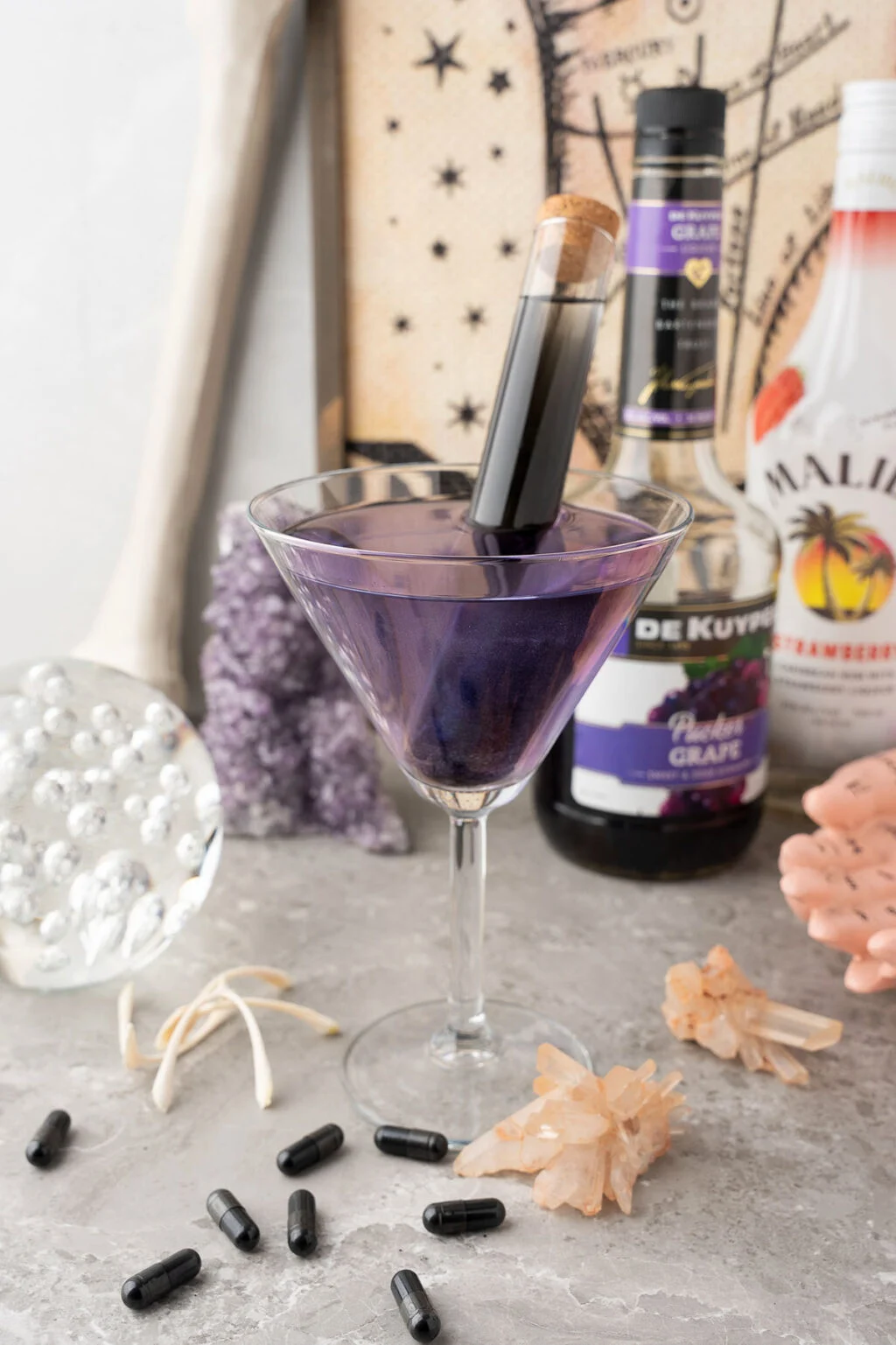 Purple alcoholic beverage in a glass with a vial inside with decorations and ingredients in the background.