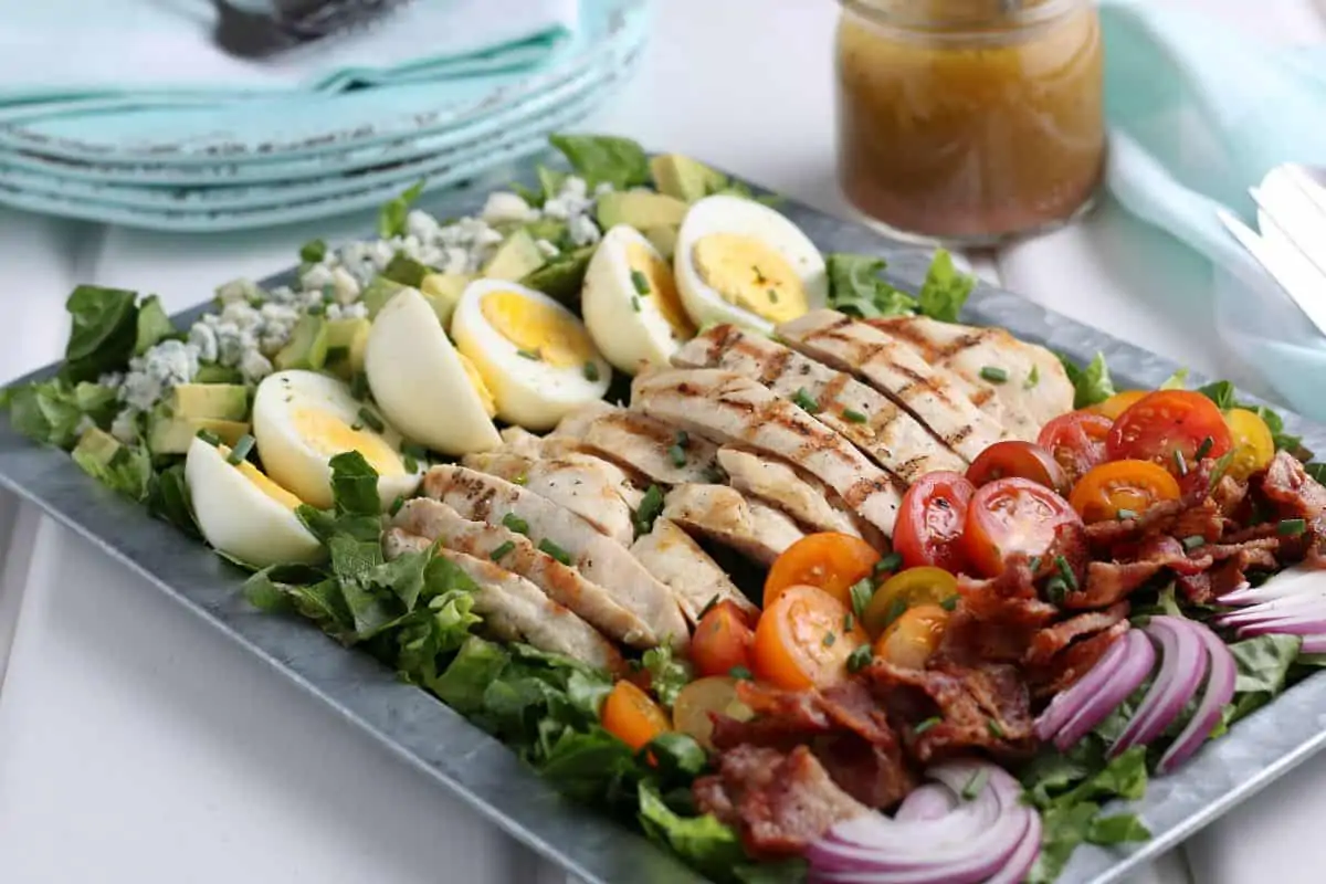 Cobb salad including eggs, chicken, tomato, bacon, onion, lettuce and blue cheese on a platter with blue plates in the background.