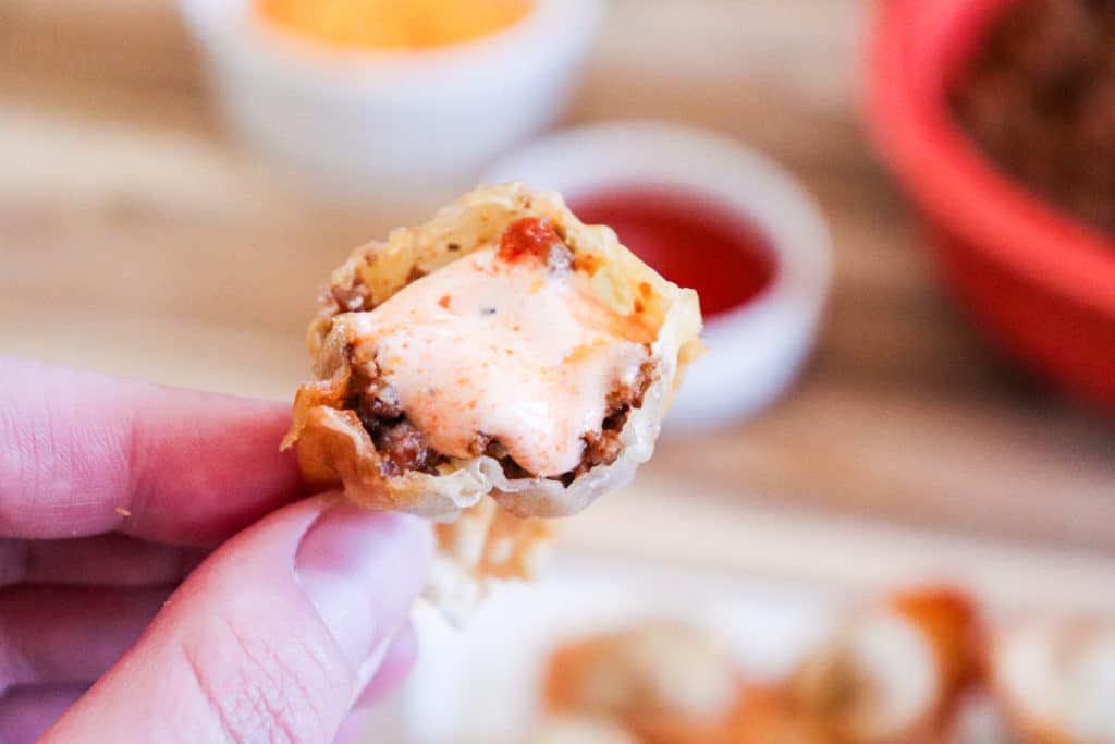 Enchilada wontons in hand with sauce in bowls in the background.