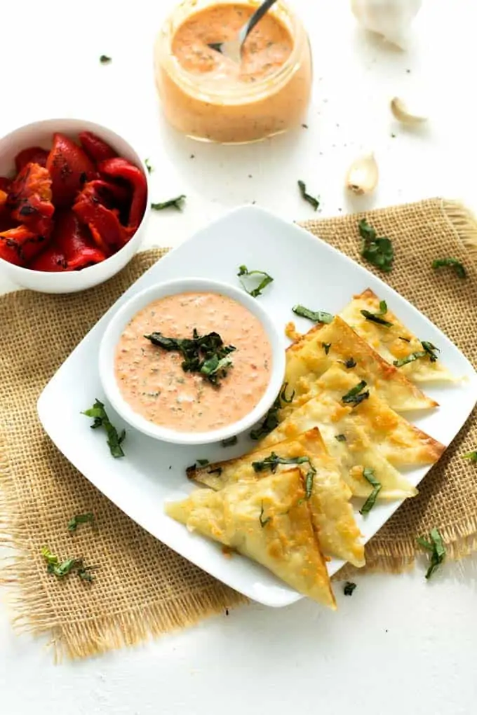 Goat cheese wontons on a white platter on top of tan fabric with roasted red peppers and dipping sauce in the background.