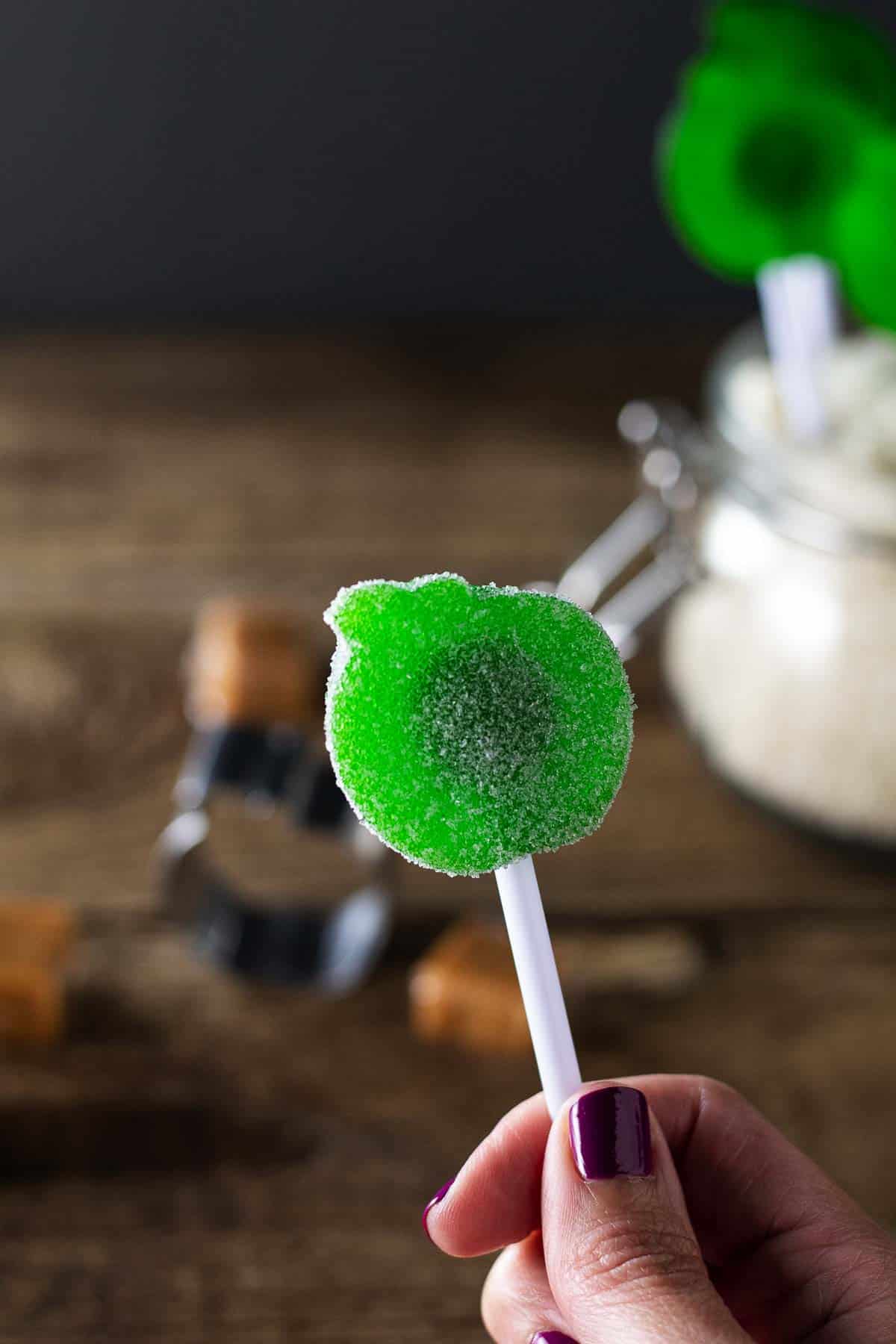 Caramel apple gummi sucker dipped in citric acid/sugar mixture held in hand with caramels and apple shape cookie cutter with wooden background.