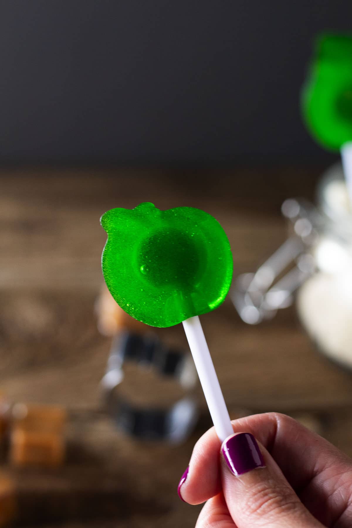 Green apple caramel gummi pop in hand with caramel candies, apple cookie cutter, and wooden background.