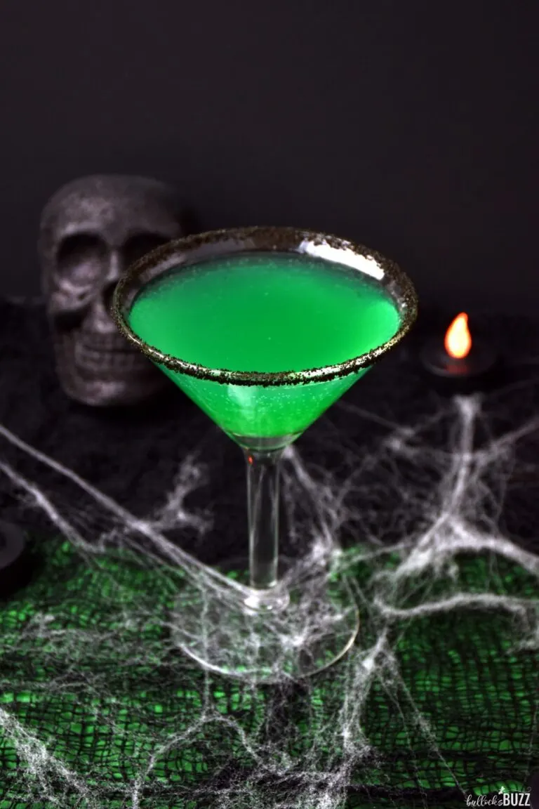 Green goblin drink in a glass rimmed with black sanding sugar and spider web, skull, and candle props in the background.