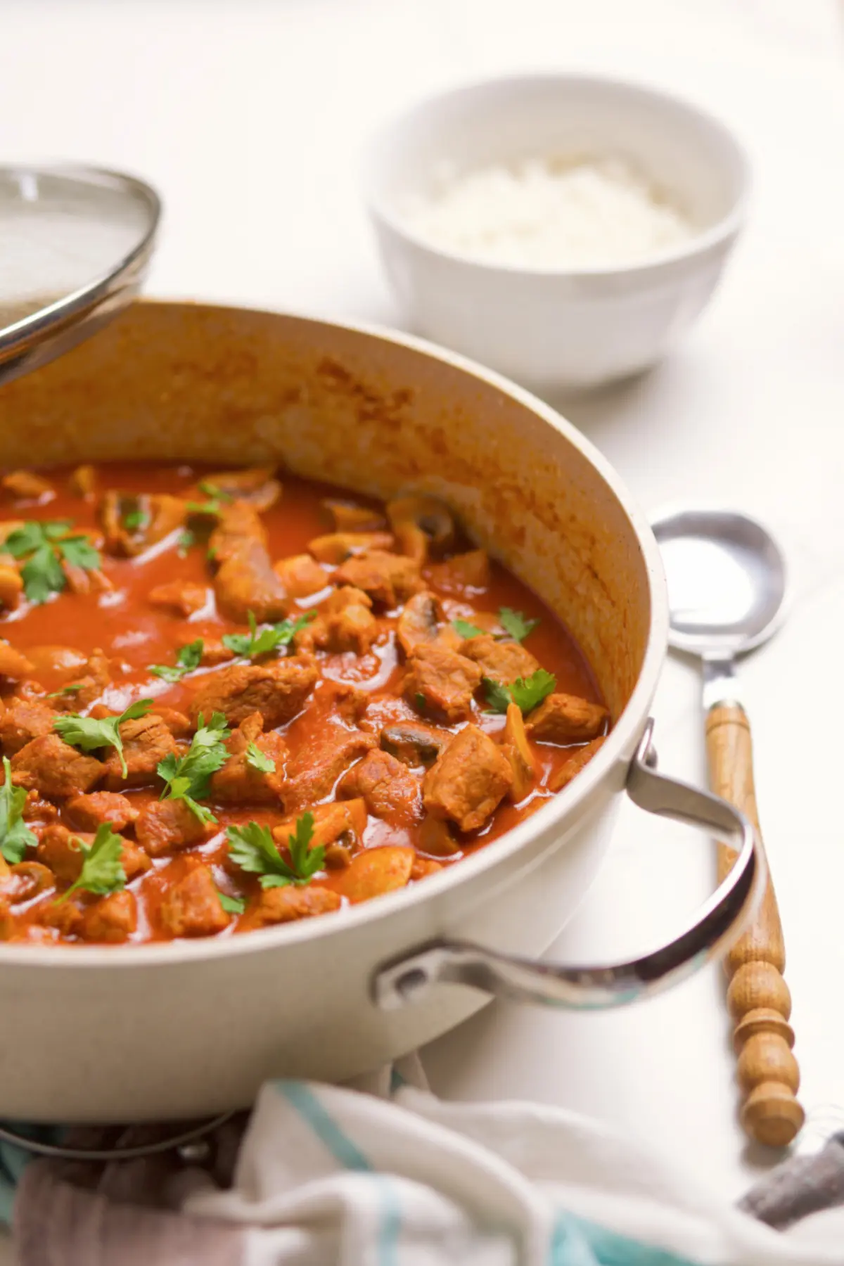 Beef goulash with beef and mushrooms in a pan with wooden spoon and bowl in rear of image.