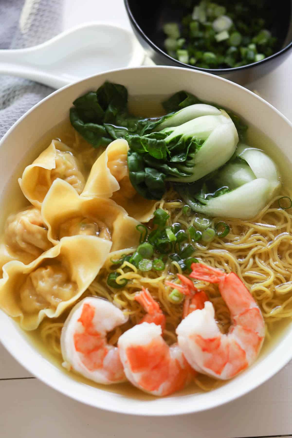 Wonton noodle soup with wontons, baby bok choy, shrimp, egg noodles, and green onions, and broth in a white bowl.