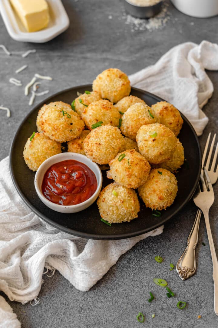 Potato croquettes on a plate with small bowl of dipping sauce with various props in background.