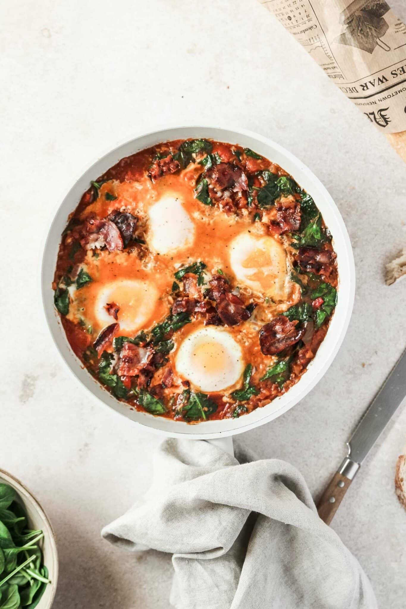 BLT shakshuka in white bowl with ingredients, knife, towel, on a light grey background.