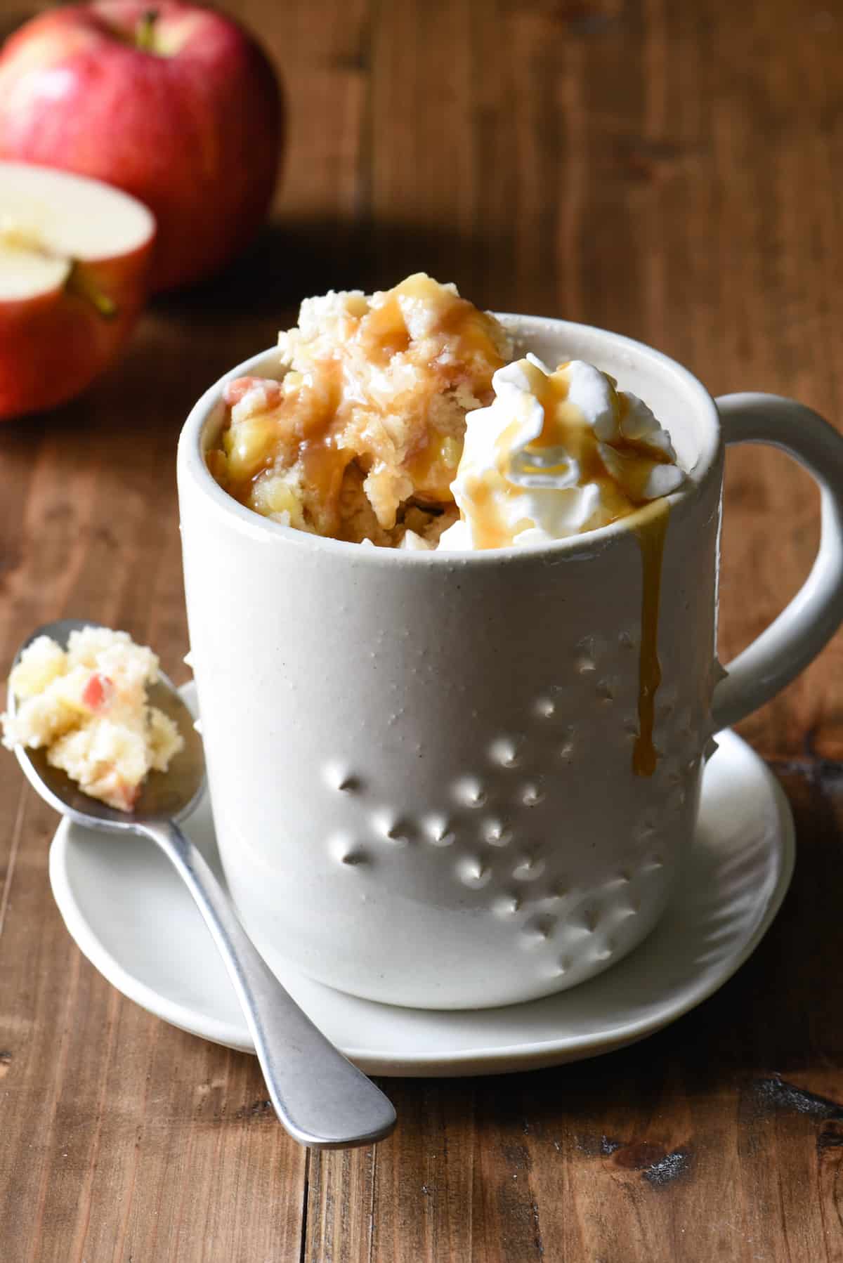 Caramel apple cake with whipped cream and caramel in a white mug with apples in background.