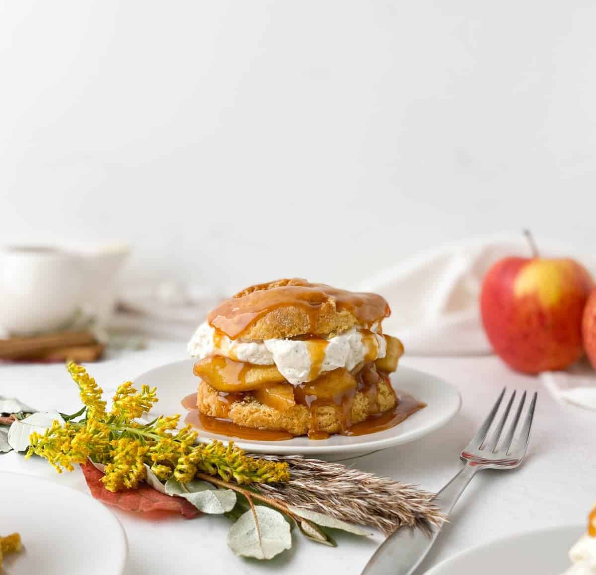 Caramel apple shortcake on a white plate with flowers, apples, and fork in background.