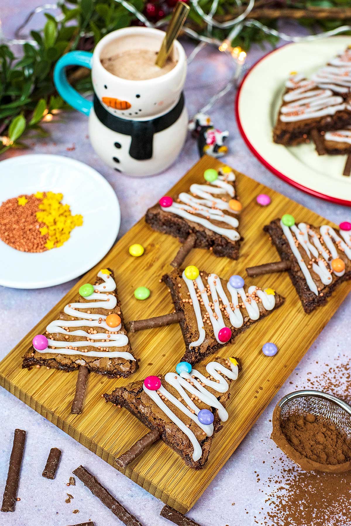 Christmas tree shaped brownies on a brown cutting board with hot chocolate in a mug and ingredients in the background.