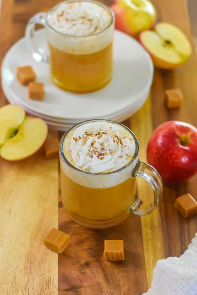 Bourbon apple cider in a clear mug with whipped cream and cinnamon on top with caramel candies and apples in background.