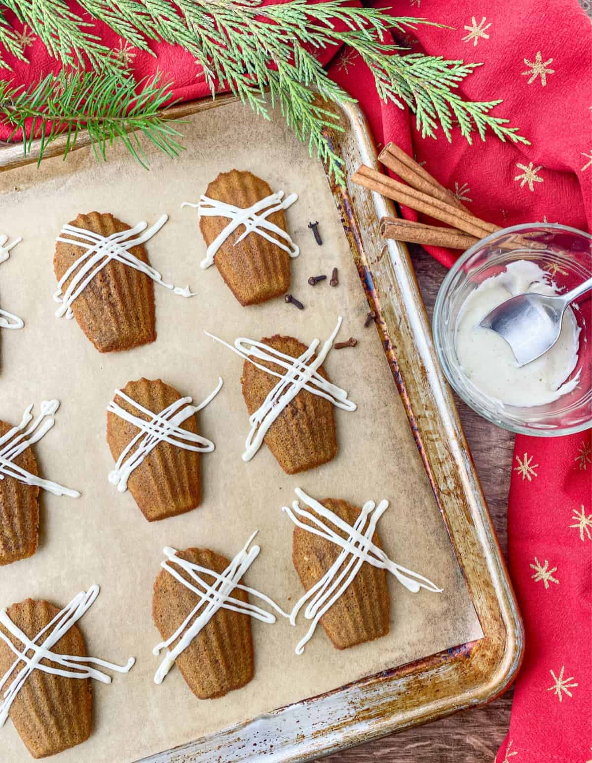 Gingerbread madeleines on a cookie sheet with vanilla bean glaze with more glaze in a small clear bowl and cinnamon sticks in the background.