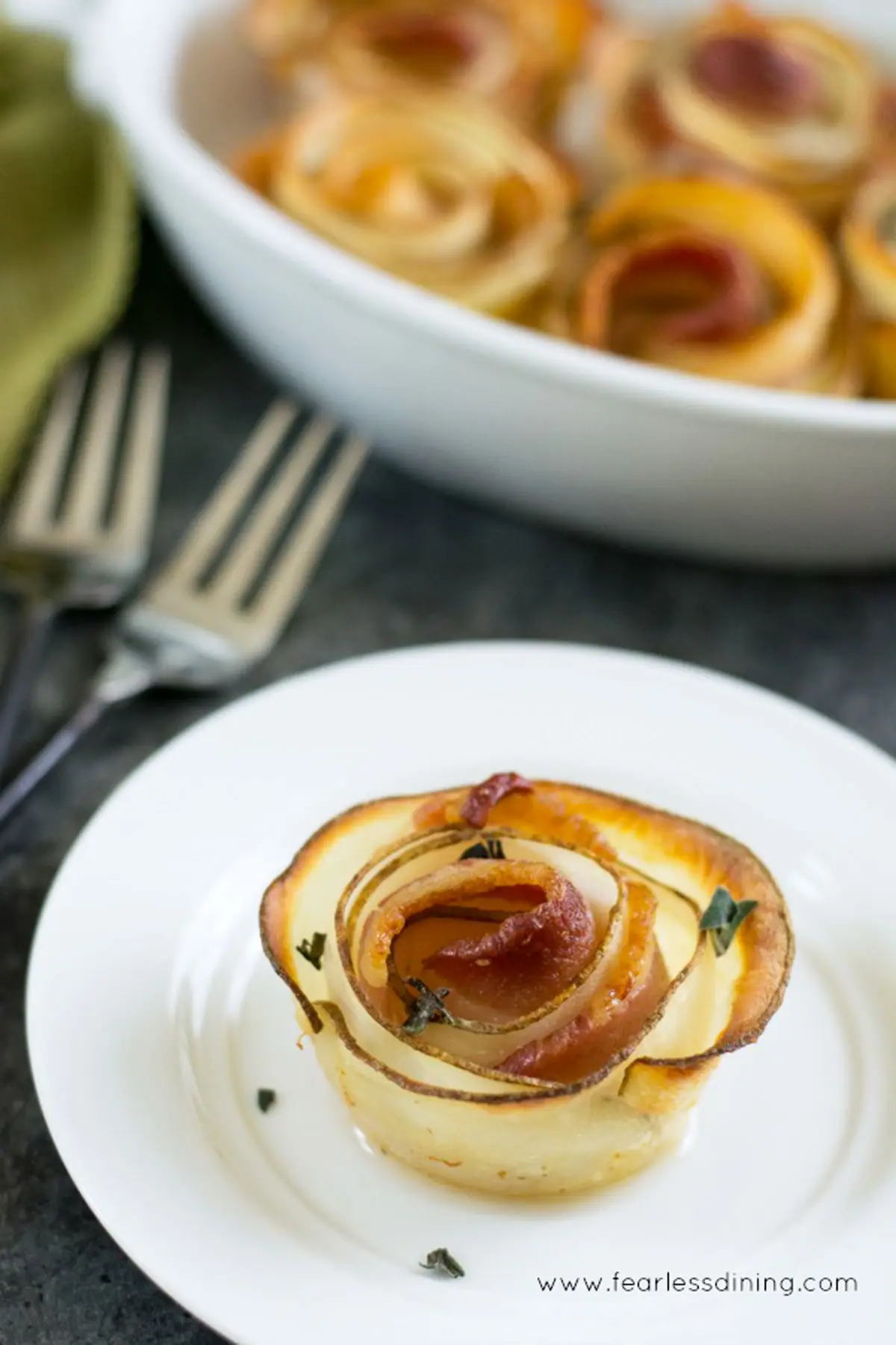 Potato-bacon rose on white plate with more in a casserole dish in background.