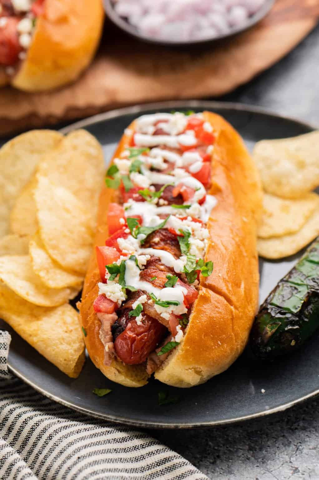 Sonoran hot dog with chips on plate with grey background.