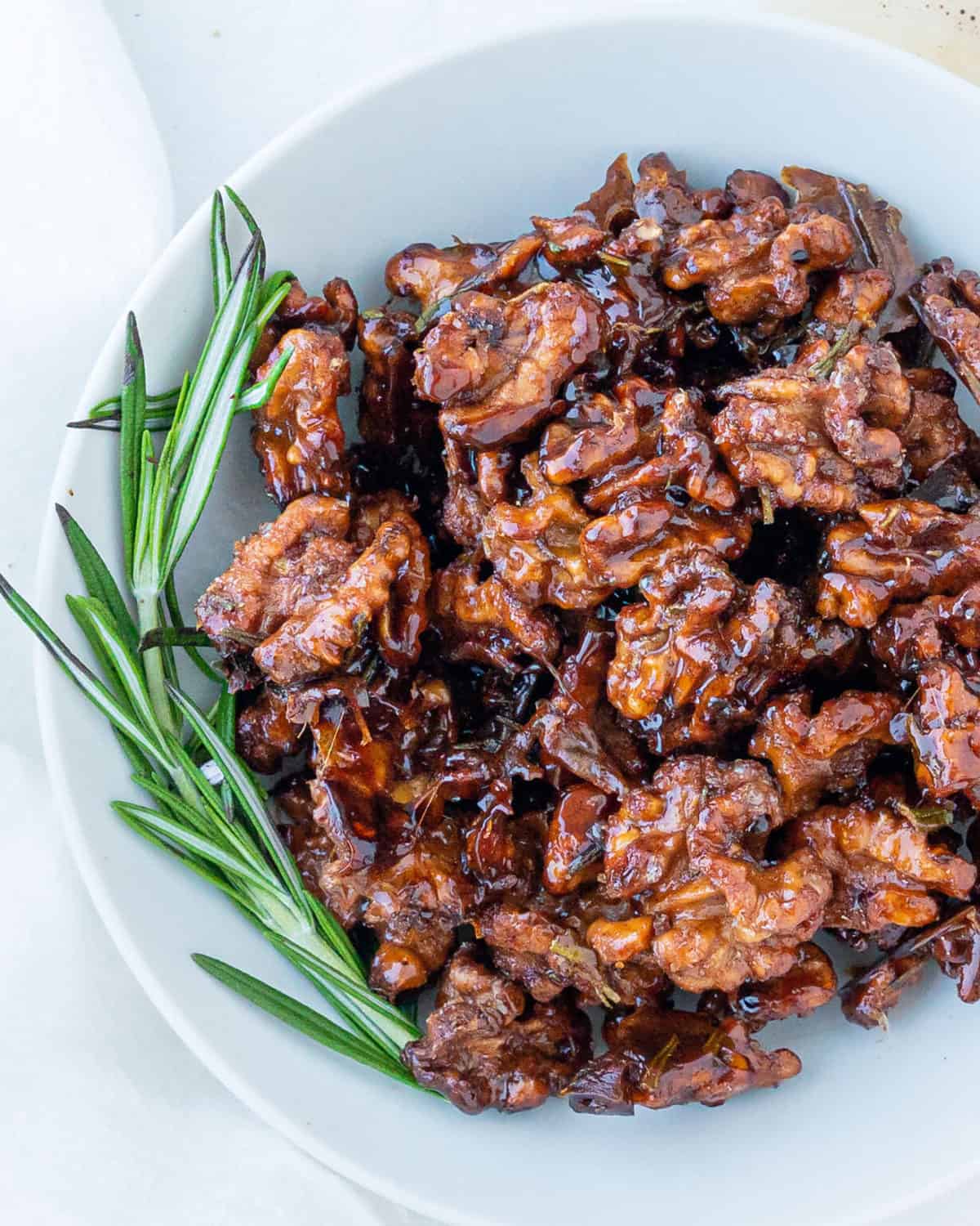 Sweet rosemary walnuts in a white bowl with rosemary sprig.