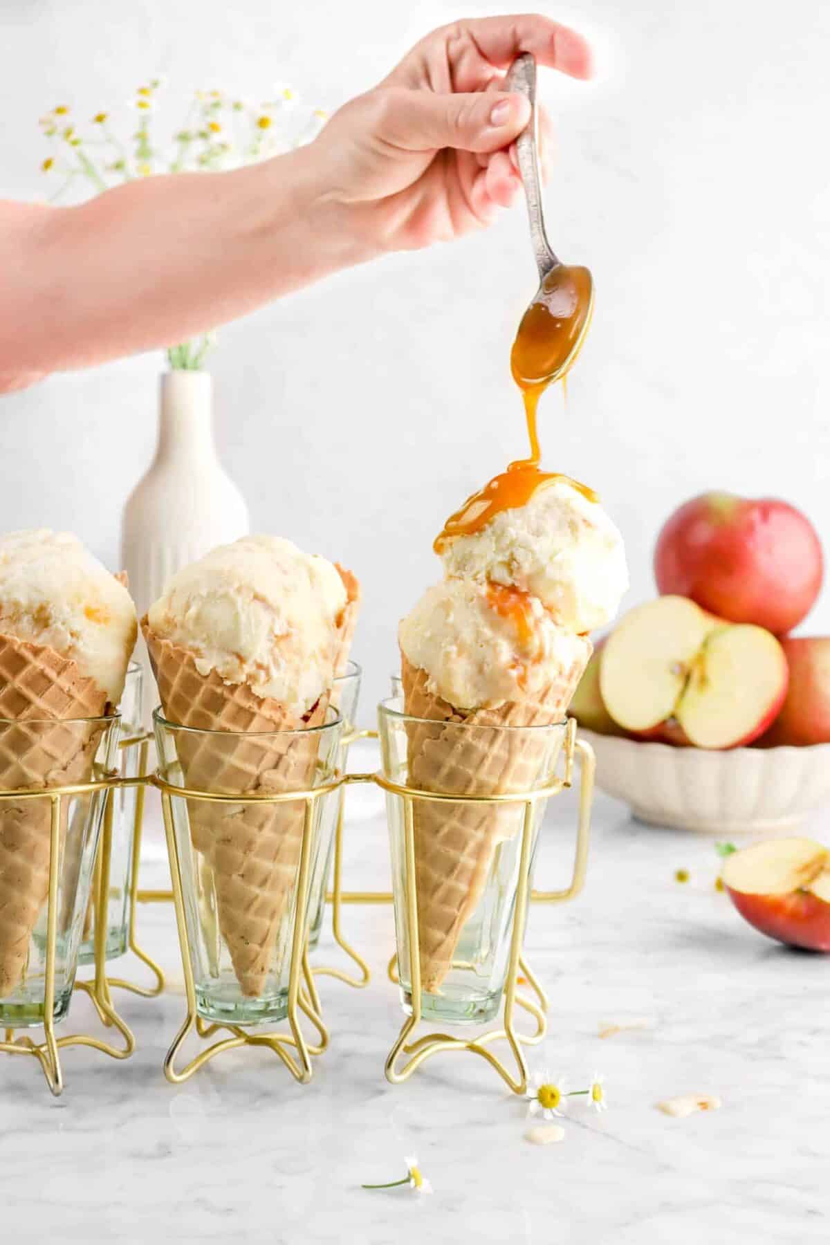 Hand drizzling caramel on ice cream cone with apples in background.