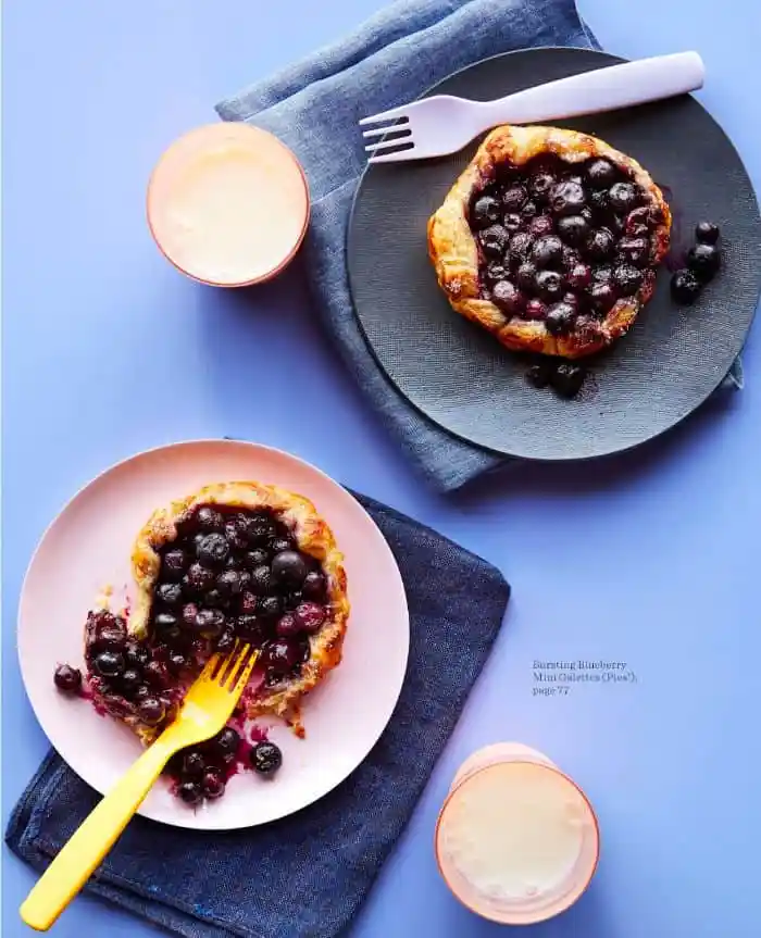 Two Blueberry galette's on pink and blue plates with cups of milk, napkins, and forks.