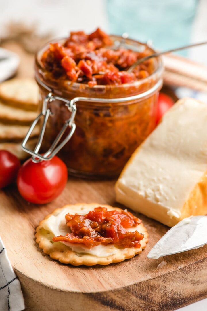 Cracker with cheese and tomato bacon jam with various food props in the background.