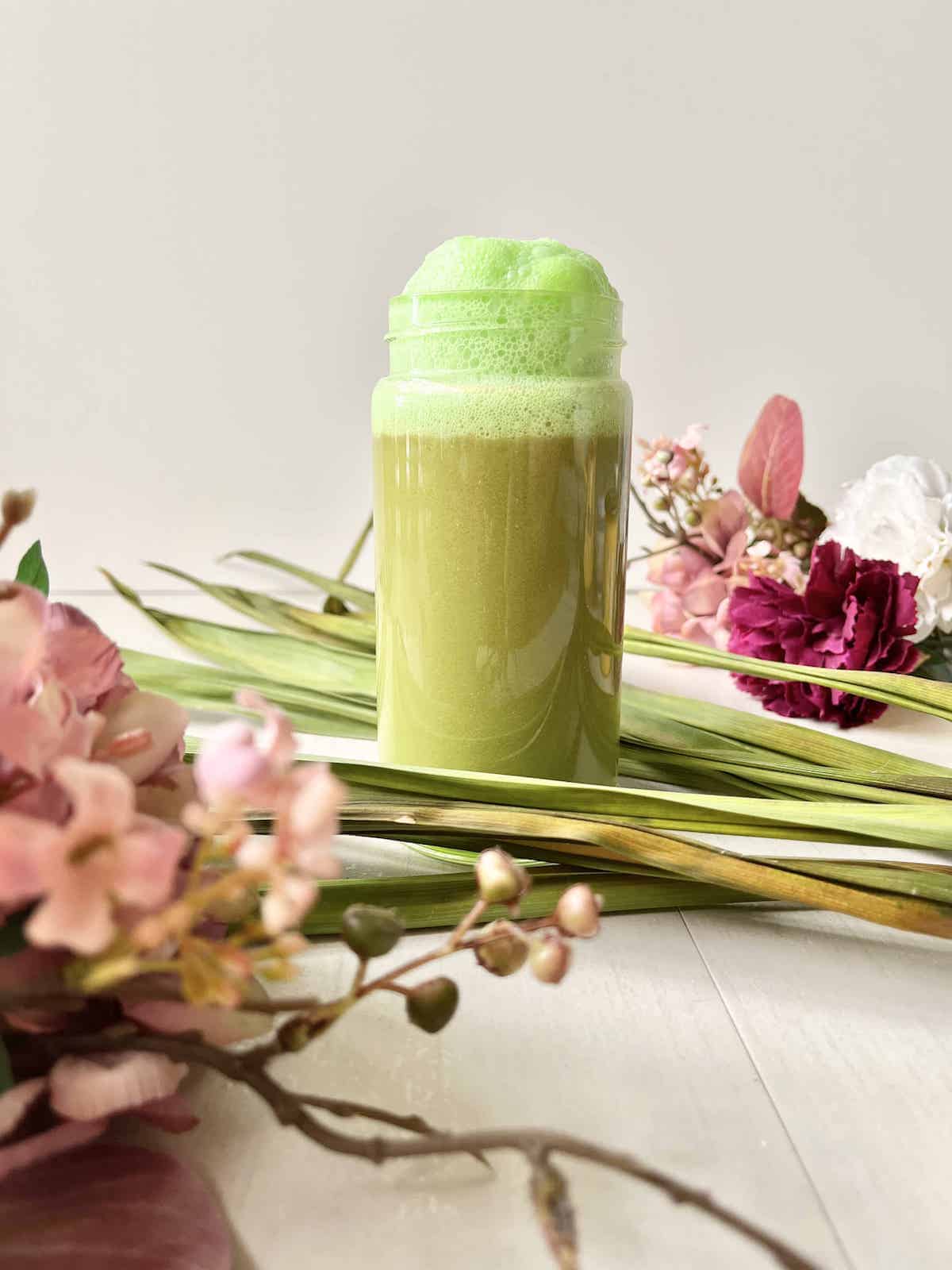 Pandan latte with green foam in container with leaves and flowers in background.