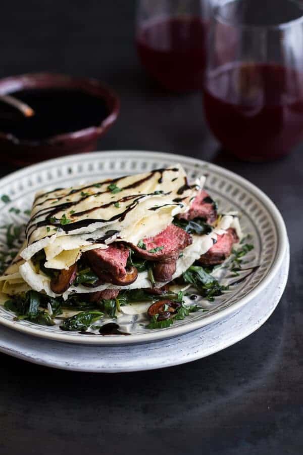 Steak, spinach, and mushroom crepes with balsamic glaze on a decorative plate with wine glasses in background.