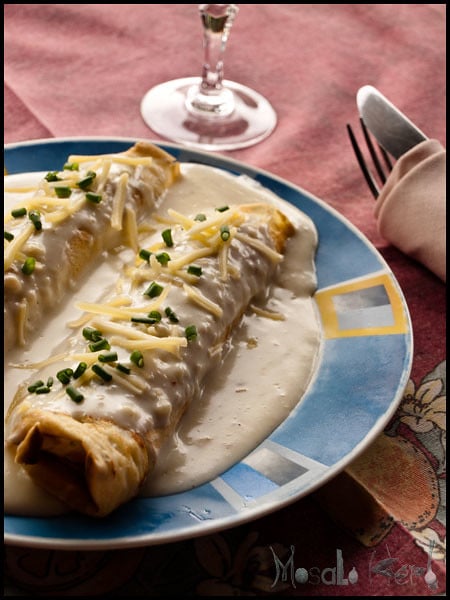 Savory stuffed crepes with white sauce, cheese, and chives on a multi color plate.