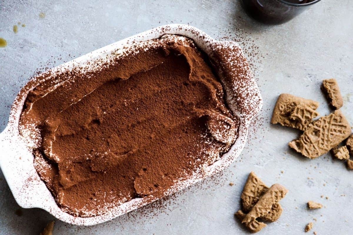 Tiramisu made with speculaas cookies in dish with broken speculaas cookies in background.