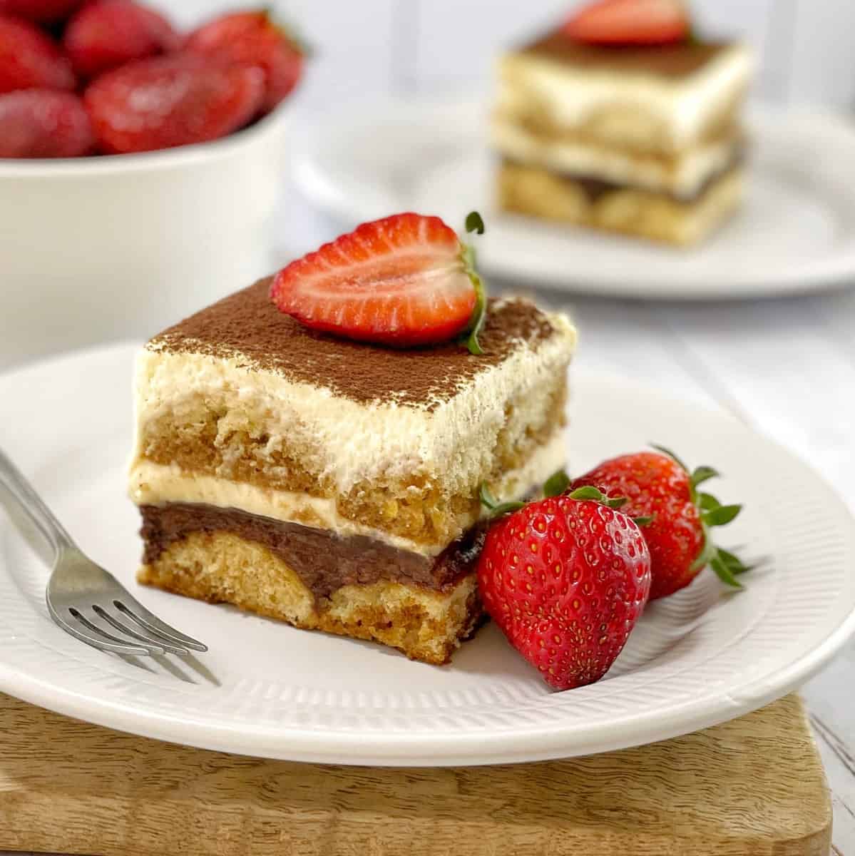 Serving of tiramisu with layer of Nutella on a white plate with another serving and strawberries in the background.