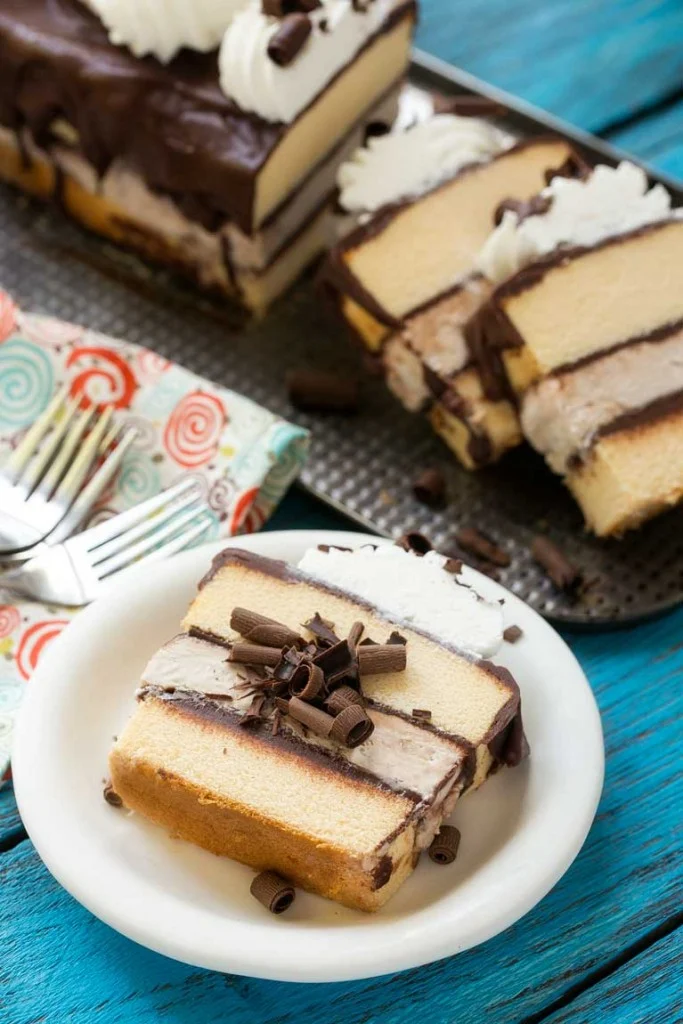 Chocolate covered coffee ice cream cake slice on white plate with more in background on blue wooden board.