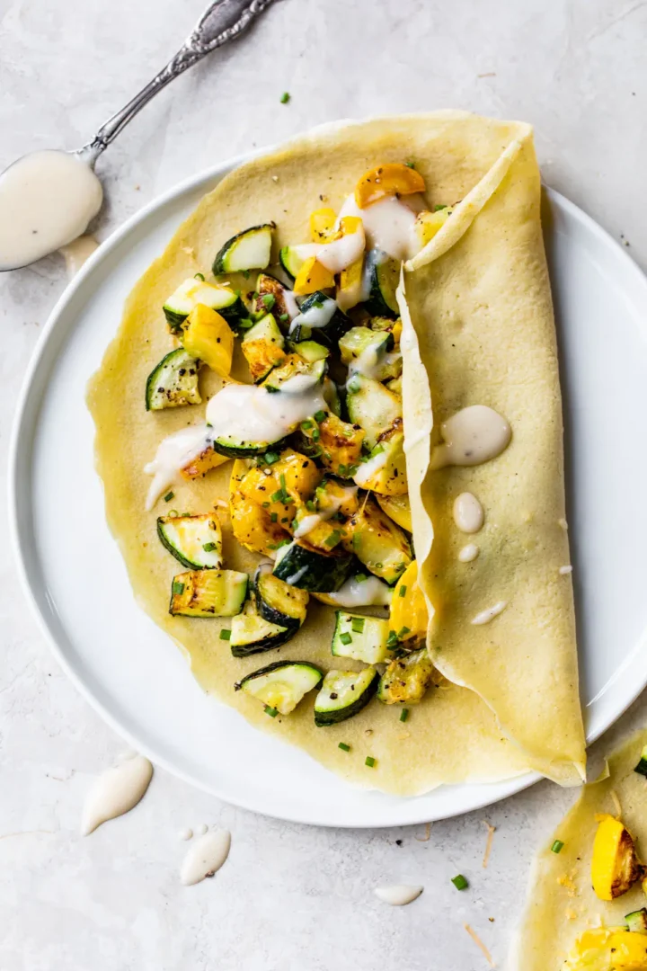 Summer squash filled crepe with bechamel sauce on a white plate.