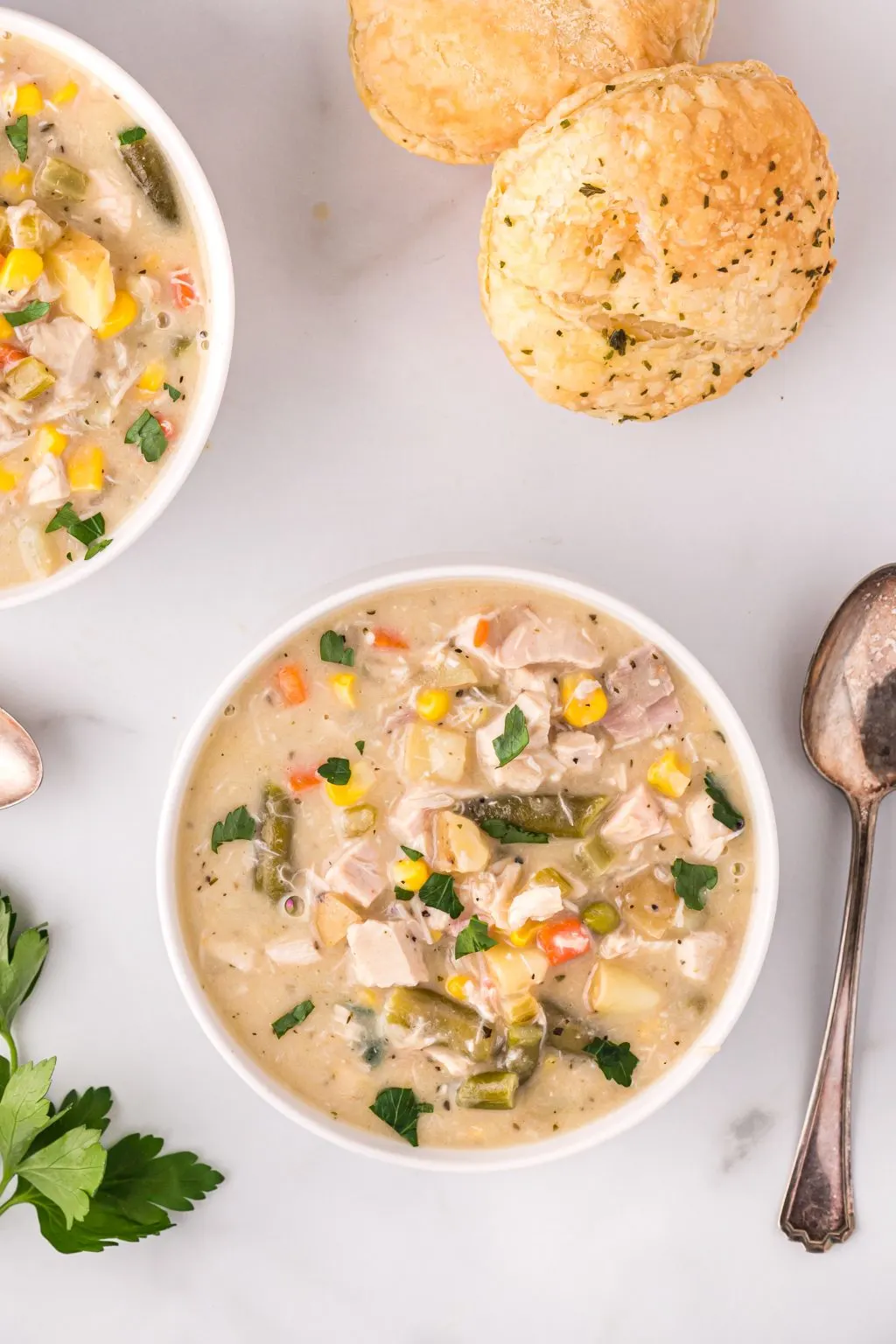 Chicken pot pie soup served in white bowls with puff pastry biscuits , herbs, and a spoon in scene.