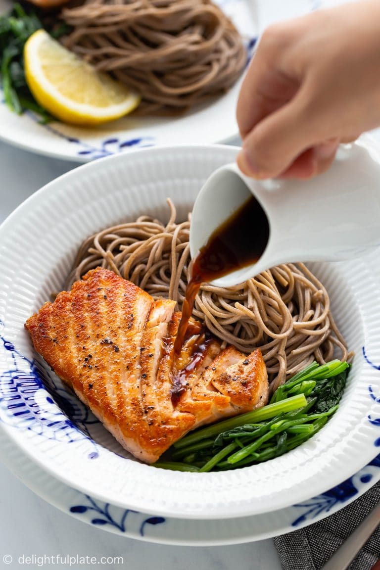 Hand pouring ginger soy dressing over salmon and soba noodles.