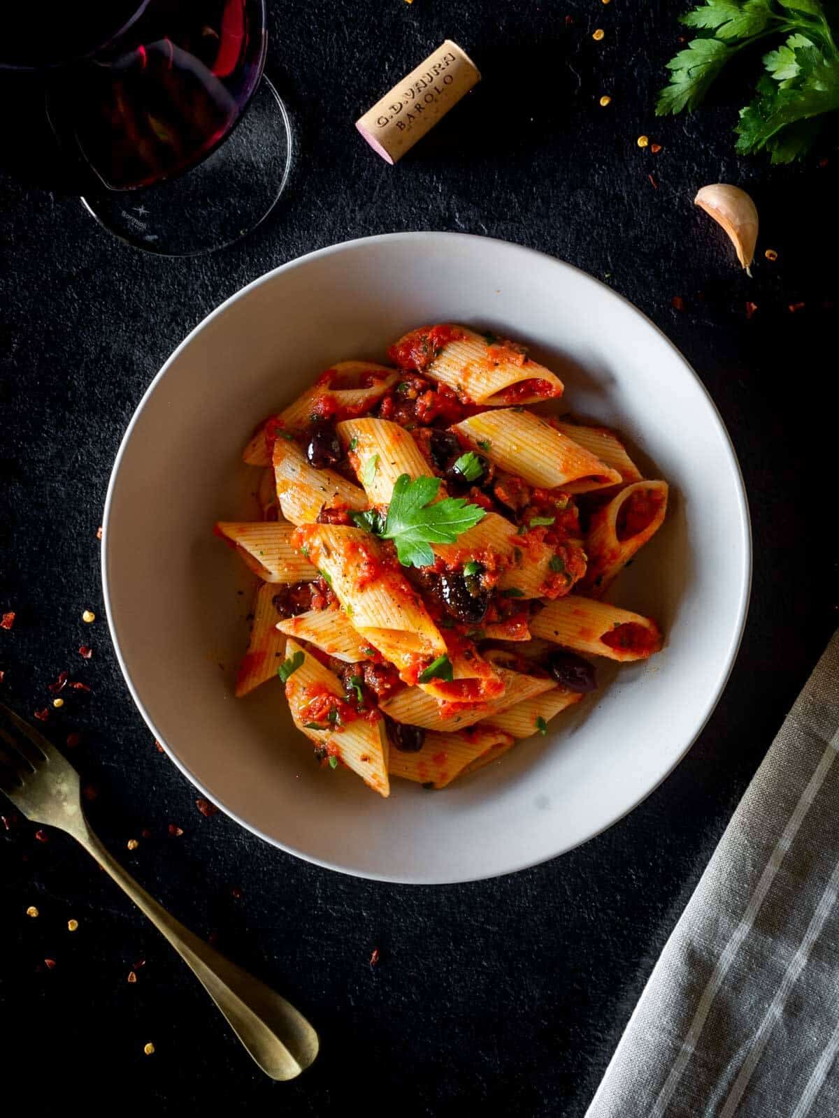 Pasta puttanesca in a white bowl with garlic, glass of wine, and herbs in background.