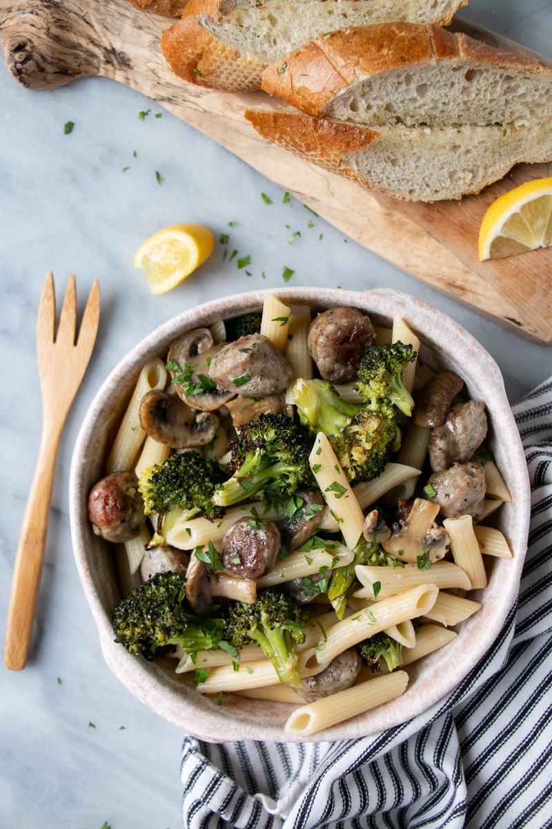 Roasted sausage and broccoli pasta in a bowl with bread, lemon wedges, and fork surrounding it.