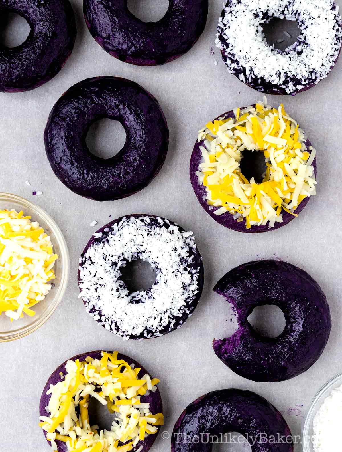 Baked purple ube donuts topped with coconut flakes and others topped with shredded cheese.