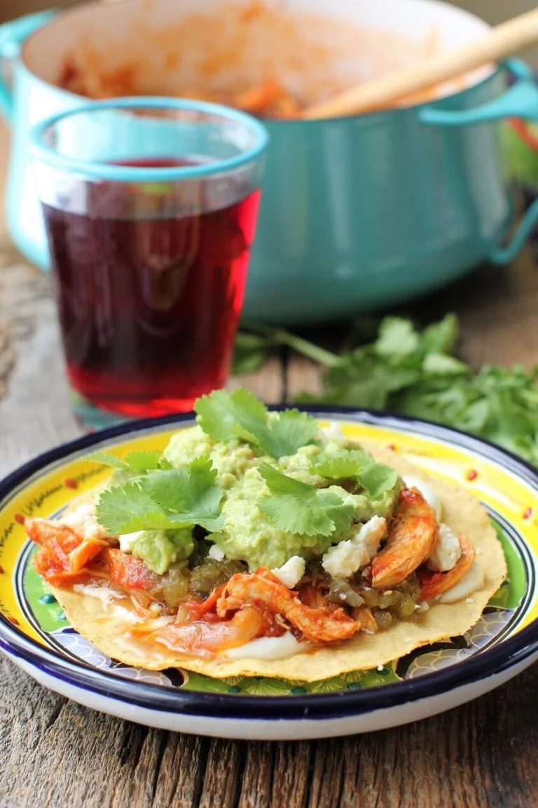 Chicken tinga tostada on a decorative plate pile high with toppings with a beverage in the background.