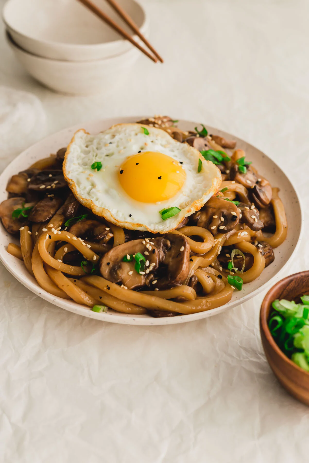 Mushroom udon noodles with egg on a plate with bowls and chopsticks in the background.