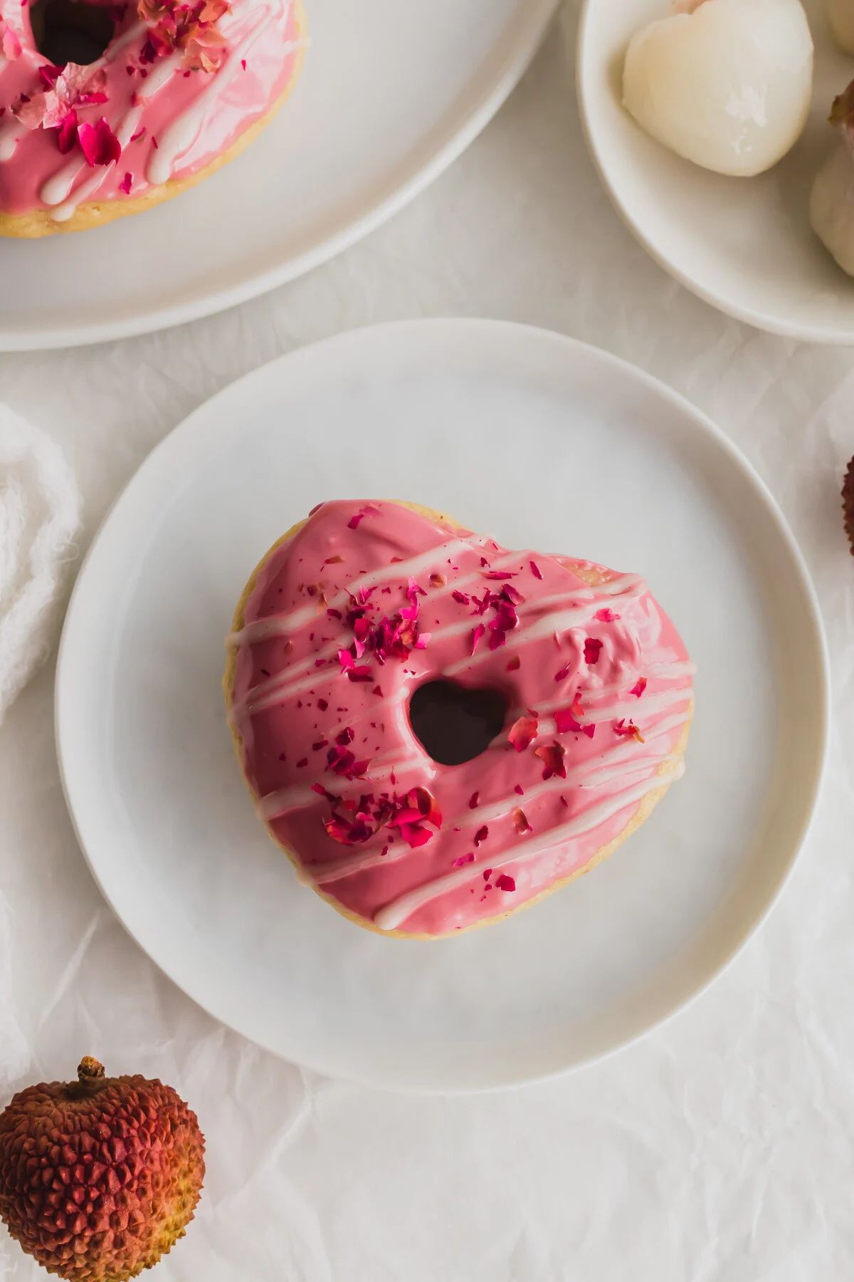 Heart shaped lychee donut with pink glaze and edible rose petals on a white plate.