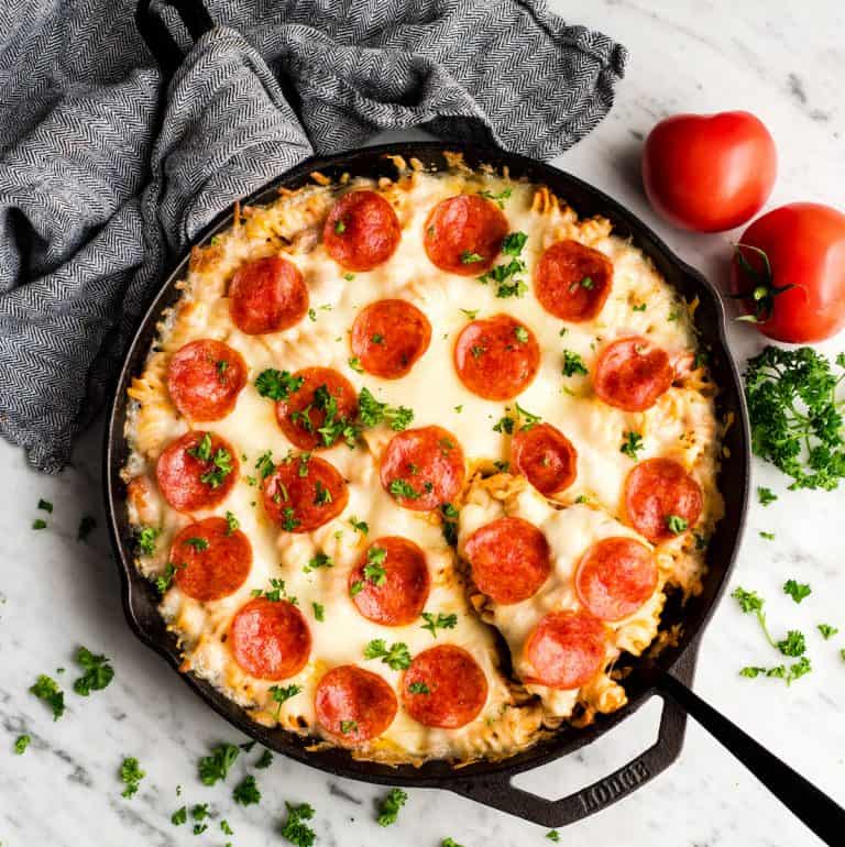 Pizza casserole in a cast iron pan with tomatoes, parsley and a dish towel in the background.