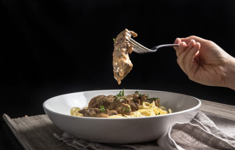 Hand holding fork with chunk of beef with more beef stroganoff under it.