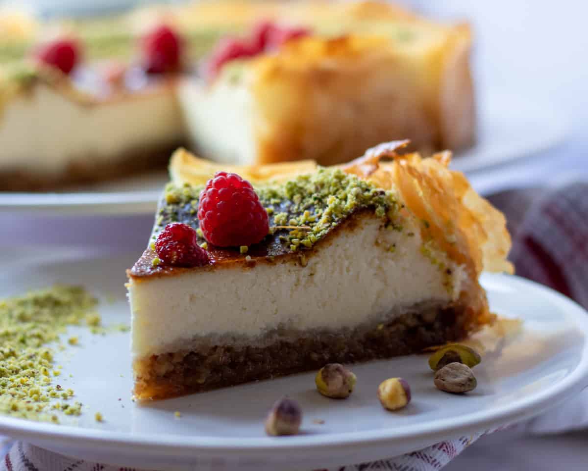 Baklava cake slice topped with pulverized pistachios and raspberries on a white plate.