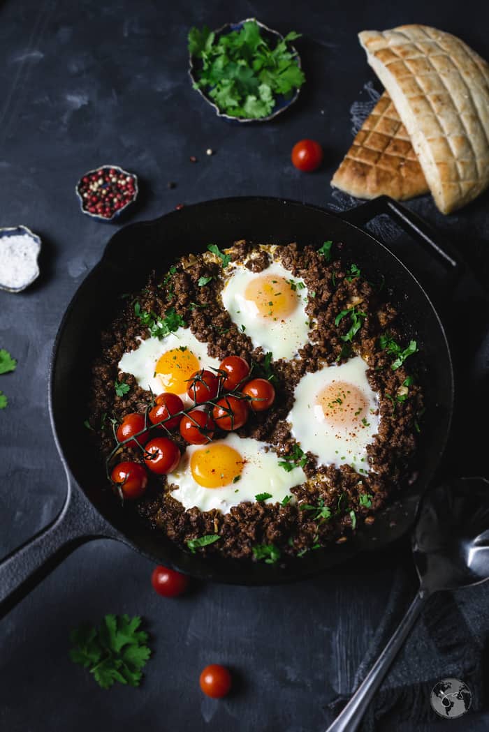 Easy minced meat and poached eggs in a cast iron pan with small tomatoes and bread and herbs in the background.