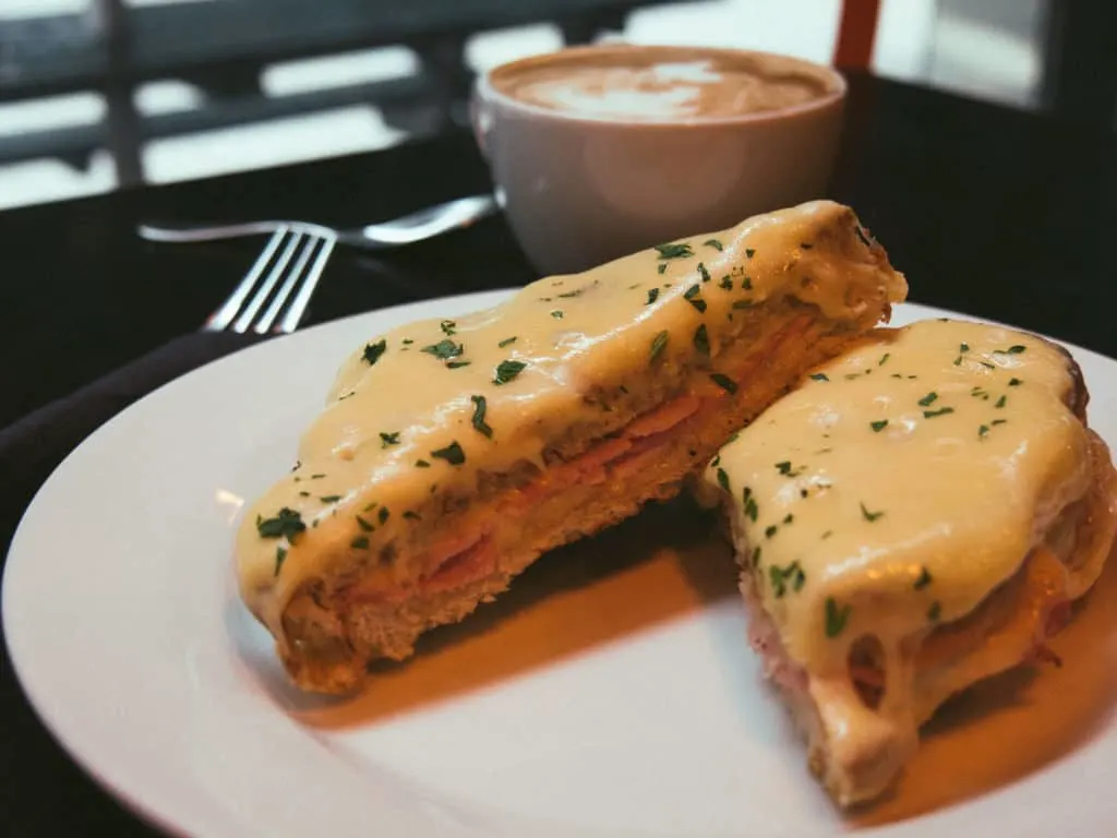 French Croque Monsieur topped with cheese and bechamel sauce on a white plate with more sauce in the background.