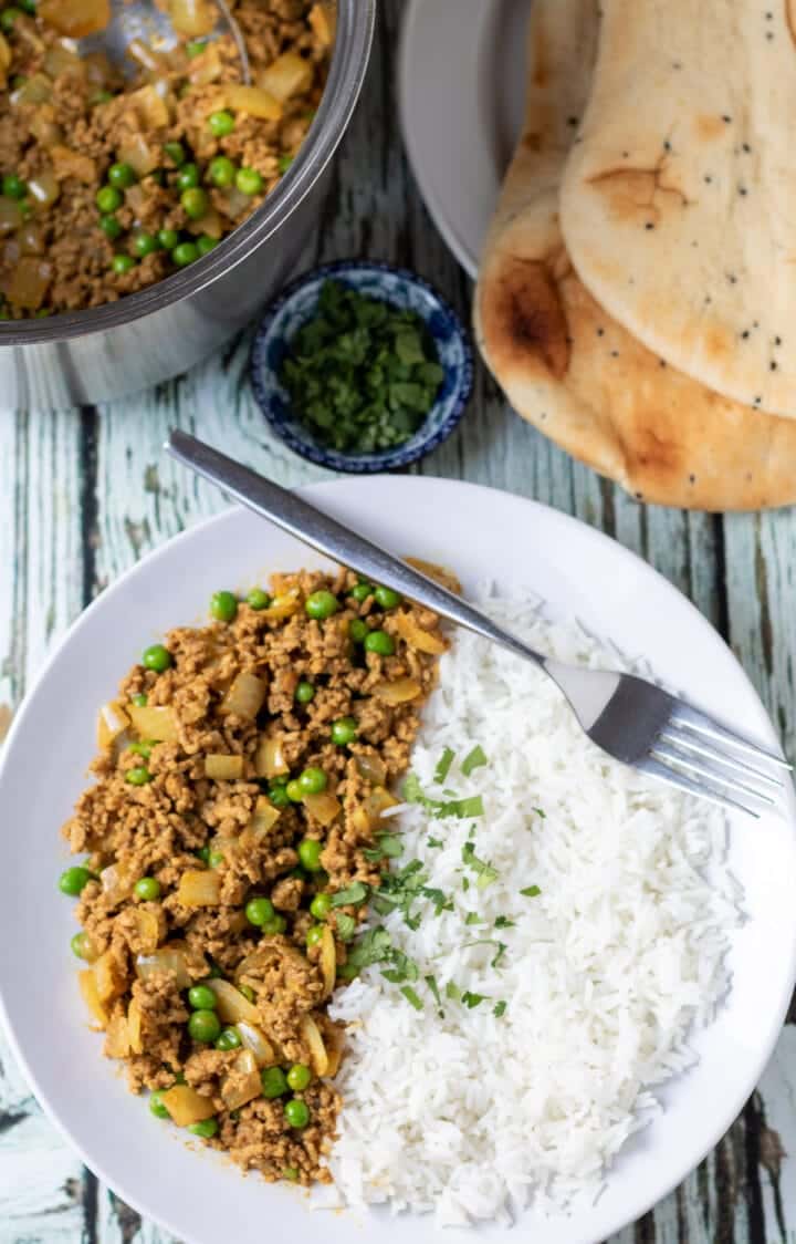 Ground lamb curry on a white plate with rice and fork with bread and more curry in the background.