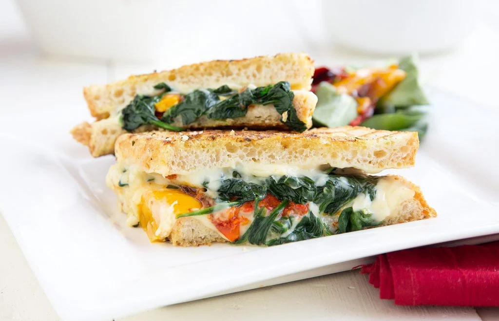 Italian grilled cheese with roasted peppers and spinach cut in half on a square white plate.