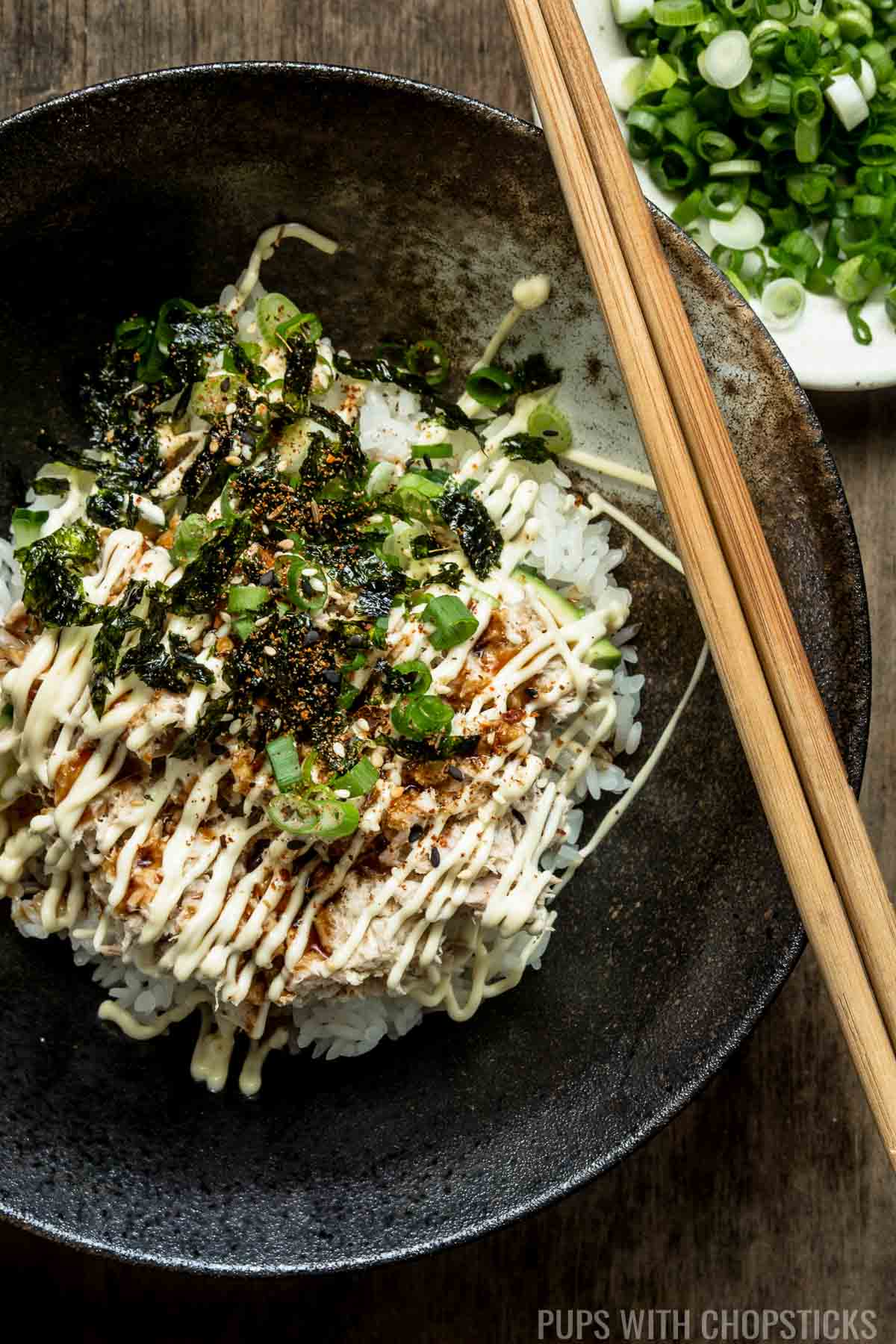 Korean tuna rice bowl in a dark bowl with a green onions and chopsticks in the background.